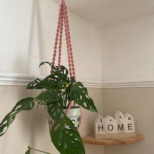Könnte beinhalten: Ein rosa Makramee-Pflanzenhänger mit einem Holzring an der Oberseite, der eine Topfpflanze mit großen, grünen Blättern hält. Die Pflanze hängt in einer Ecke eines Raumes, mit einem Holzregal darunter. Das Regal hat ein Holzschild mit der Aufschrift "HOME" darauf.
