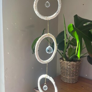 May include: A three-tiered, macrame-style wall hanging with white rope rings and clear crystal pendants. The rings are suspended by gold-colored string. A potted plant is in the background, adding a touch of nature to the decor.