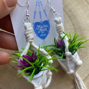 May include: A pair of white macrame earrings with small white pots and green and purple plants. The earrings are attached to silver hooks. The earrings have a gold accent on the macrame. The earrings are labeled "Macra Made with love".