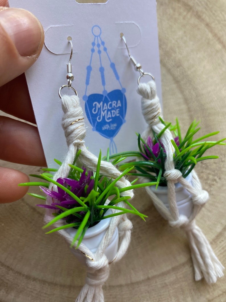 May include: A pair of white macrame earrings with small white pots and green and purple plants. The earrings are attached to silver hooks. The earrings have a gold accent on the macrame. The earrings are labeled "Macra Made with love".