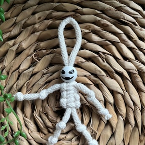 May include: A handmade white macrame skeleton figure with a black painted face. The skeleton has a loop at the top for hanging. The figure is displayed on a woven, textured background with green foliage.