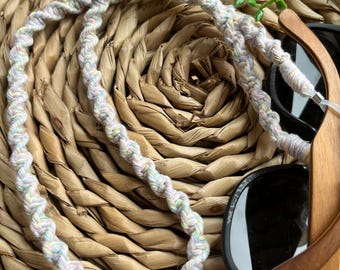 May include: A white, pink, and yellow braided cord with a twisted design. The cord is looped around a pair of wooden sunglasses with black lenses.