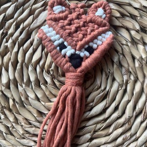 May include: A handmade macrame fox keychain in shades of peach, white, and black. The fox design features a detailed face with a tassel tail and a silver keyring. The keychain is made of woven cotton cord.