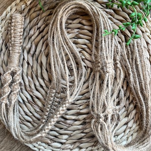 May include: Two macrame plant hangers made from natural jute rope. The hangers are displayed on a woven wicker surface.