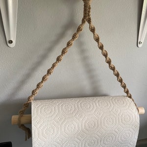 May include: A macrame paper towel holder with a natural wood dowel and ring. The holder is made of tan-colored rope and is designed to hang on a wall. The paper towel roll is white with a repeating pattern.