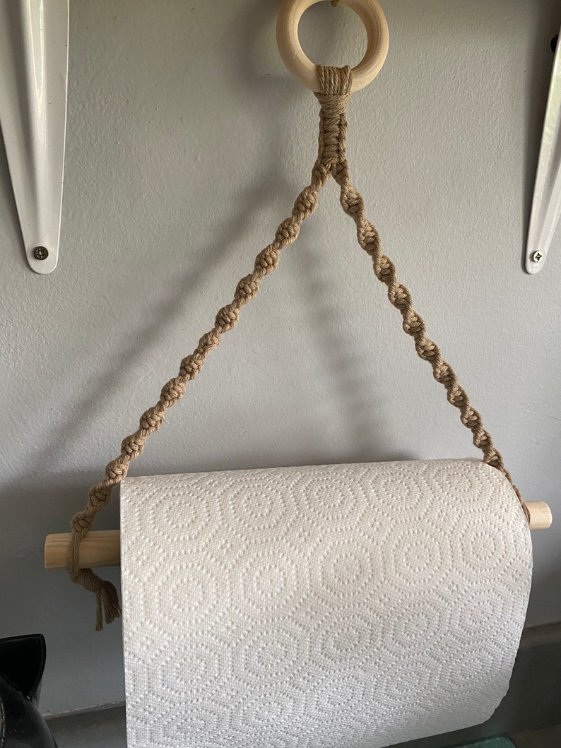 May include: A macrame paper towel holder with a natural wood dowel and ring. The holder is made of tan-colored rope and is designed to hang on a wall. The paper towel roll is white with a repeating pattern.