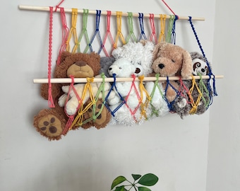 Puede incluir: Una colorida pared colgante de macramé con una varilla de madera y un arcoíris de hilo. La pared colgante está llena de animales de peluche, incluyendo un oso marrón, un oso blanco, un perro marrón y un perezoso gris.