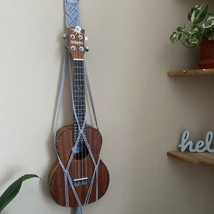 May include: A gray macrame wall hanging with a wooden ring at the top, holding a brown ukulele with the word "Octopus" on the headstock. The ukulele is hanging on a white wall.