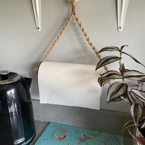 May include: A macrame paper towel holder with a wooden dowel and a natural rope design. The holder is suspended from a wooden ring. A roll of white paper towels is in the holder. The holder is hanging in a kitchen.