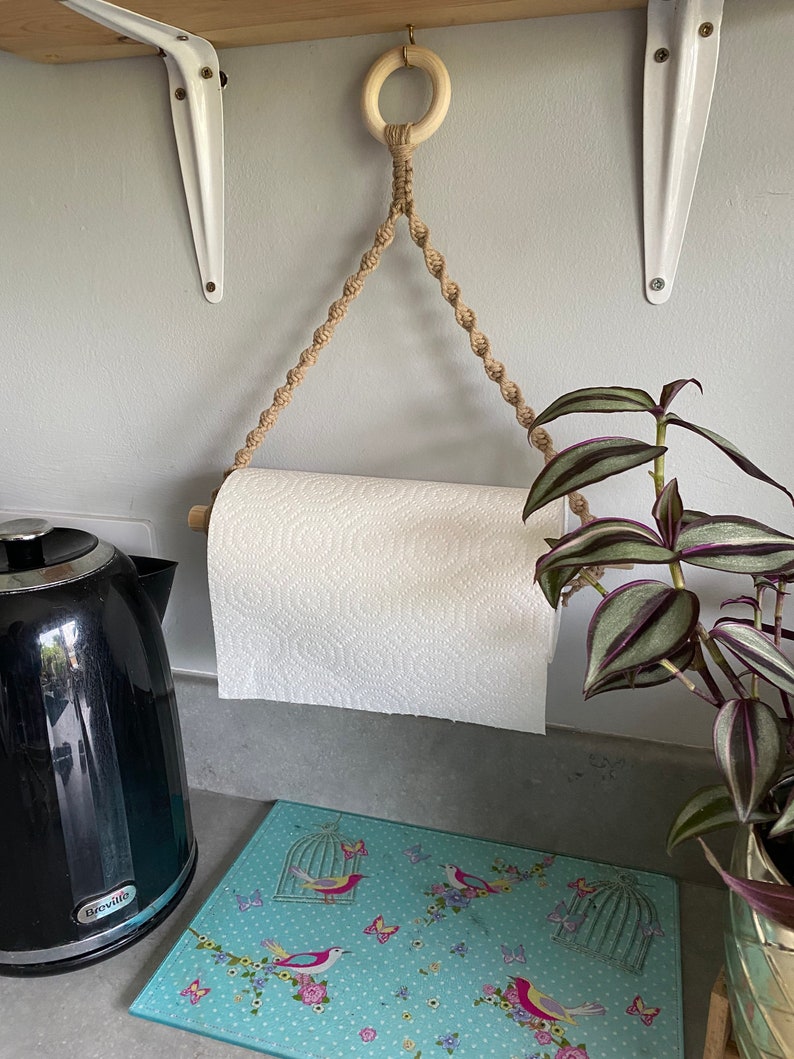May include: A macrame paper towel holder with a wooden dowel and a natural rope design. The holder is suspended from a wooden ring. A roll of white paper towels is in the holder. The holder is hanging in a kitchen.