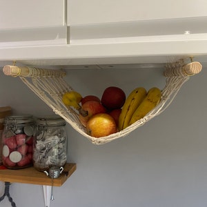 A white macrame hammock hanging from a white cabinet. The hammock is filled with bananas and red apples.