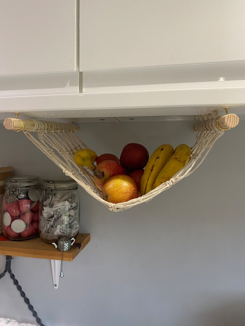 A white macrame hammock hanging from a white cabinet. The hammock is filled with bananas and red apples.
