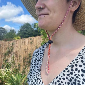 May include: A straw sun hat with a braided chin strap in shades of pink, orange, and cream. The hat is worn with a black and white patterned top. The background features a blue sky and greenery.