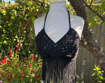 Peut inclure: Haut de bikini noir en macramé avec des franges. Le haut est présenté sur un mannequin en extérieur, avec de la verdure et des fleurs en arrière-plan. Le haut a un col en V et une bordure à franges.