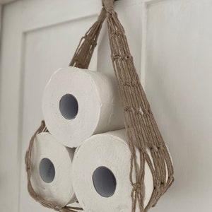 Macrame toilet paper hammock, made with taupe recycled cotton, displayed with 3 large white toilet rolls inside, hanging against a white door