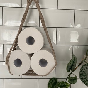 Macrame toilet paper hammock, made with taupe recycled cotton, displayed with 3 large white toilet rolls inside, hanging under a shelf against a white tiled wall with a green plant next to it
