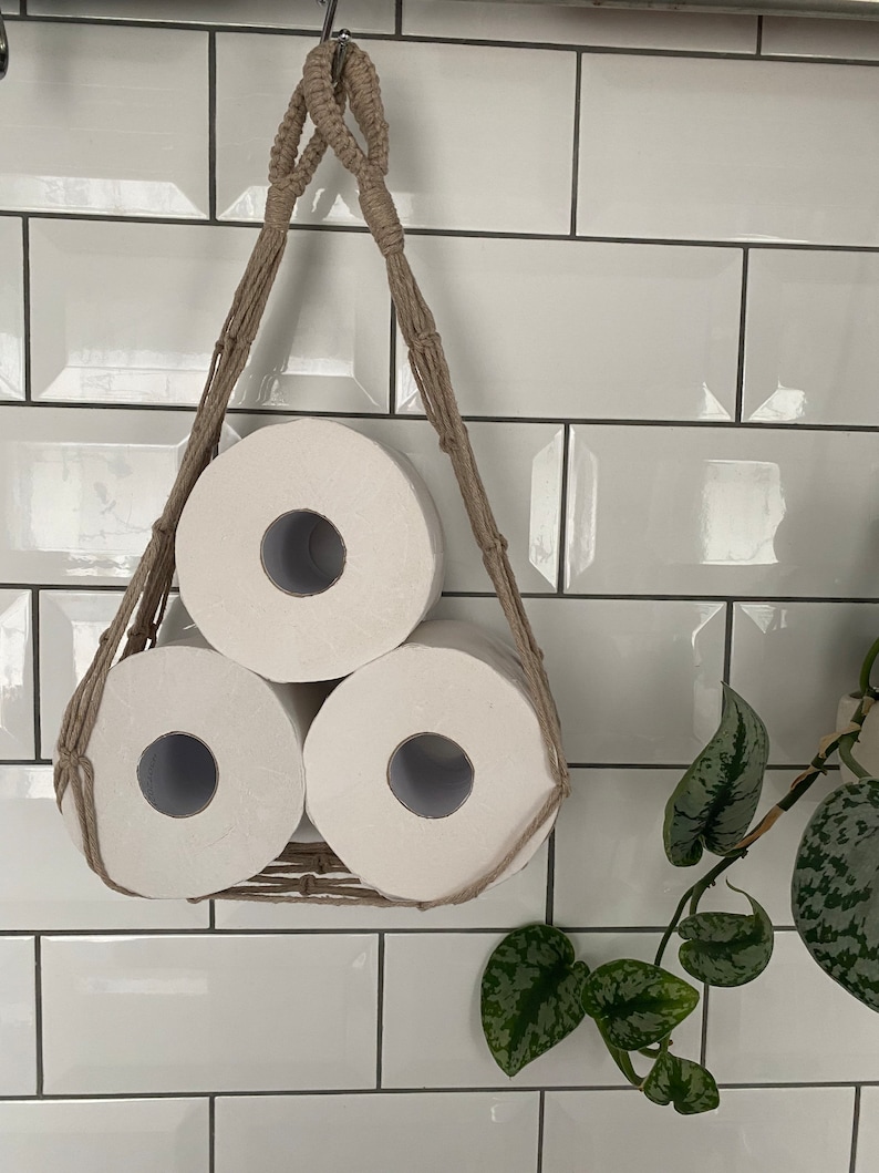 Macrame toilet paper hammock, made with taupe recycled cotton, displayed with 3 large white toilet rolls inside, hanging under a shelf against a white tiled wall with a green plant next to it