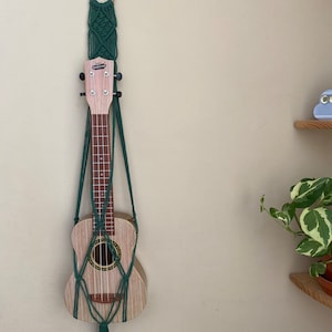 Może przedstawiać: Zielony wiszący makramowy z drewnianym pierścieniem na górze, trzymający ukulele. Ukulele jest z jasnego drewna z ciemnym palisandrem.