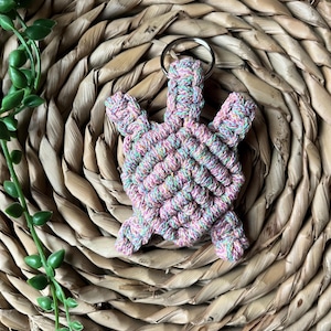 May include: A colorful, pink, green, and yellow macrame turtle keychain. The turtle is made of knotted string and has a silver key ring.