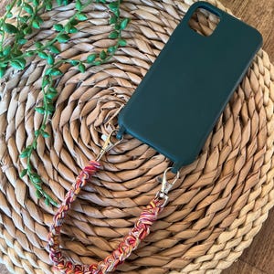 May include: A dark green phone case with a multi-coloured braided strap. The strap has red, yellow, and pink threads, attached with silver clasps. The case sits on a woven, circular mat.