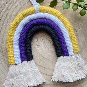 May include: A yellow, white, purple, black and white crocheted rainbow wall hanging with tassels. The rainbow is made of yarn and has a white string hanging from the top.