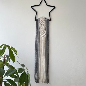 May include: A grey, white and black macrame wall hanging with a star-shaped metal frame. The macrame is made with a variety of knots and is hanging from the ceiling.