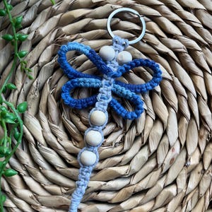 May include: A blue and gray macrame dragonfly keychain with wooden beads. The keychain is hanging on a woven wicker surface.