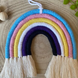 May include: A colourful macrame rainbow wall decoration. The rainbow design includes layers of blue, pink, yellow, white, purple, and black, with cream-coloured fringe. A white cord is attached for hanging. The craft is displayed on a wooden surface.