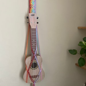 May include: A ukulele hanging on a wall with a rainbow macrame wall hanger. The ukulele is a light wood color with a dark wood sound hole. The macrame hanger is made with a variety of colors including red, orange, yellow, green, blue, and purple.