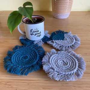 Peut inclure: Quatre dessous-de-verre en macramé faits à la main avec des franges. Deux dessous-de-verre sont gris et deux sont bleus. Les dessous-de-verre sont sur une surface en bois. Une tasse en émail blanc avec le texte "liven up your cup" est posée sur l'un des dessous-de-verre.