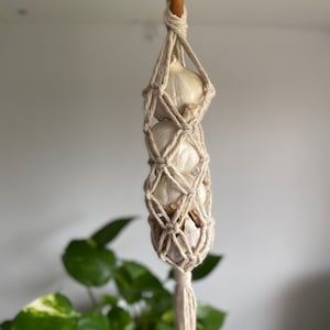 Könnte beinhalten: Eine weiße Knoblauchzehe, die in einem Makramee-Netz mit einem Holzhaken hängt. Der Knoblauch hängt in einer Küche oder einem Esszimmer.
