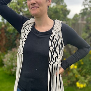 May include: A white crocheted vest with a fringe detail. The vest is worn over a black long-sleeved shirt and blue jeans.