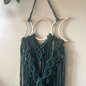 May include: A dark teal macrame wall hanging with a triple moon design. The wooden crescent moons and circular centre are connected to a woven macrame design with long, flowing fringe. The wall decor is suspended by a dark teal cord.