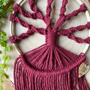 May include: A maroon macrame wall hanging in the shape of a tree with a wooden hoop and a heart-shaped wooden tag that says "You are loved".