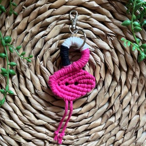 May include: Pink and black macrame flamingo keychain with a white accent. The keychain is attached to a silver key ring.