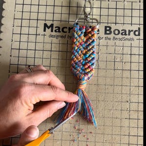 May include: A vibrant macrame keychain with a silver clasp. The keychain features braided threads in blue, orange, yellow, and red. The background shows the text "Macro Board designed by for the BeadSmith".