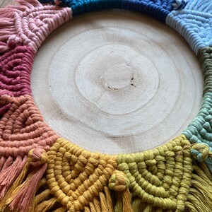 Puede incluir: Un tapiz de macram&eacute; colorido con un arco&iacute;ris de colores, incluyendo rosa, rojo, naranja, amarillo, verde, azul y morado. El macram&eacute; est&aacute; hecho con una variedad de nudos y est&aacute; terminado con borlas. El tapiz se muestra en un c&iacute;rculo de madera.