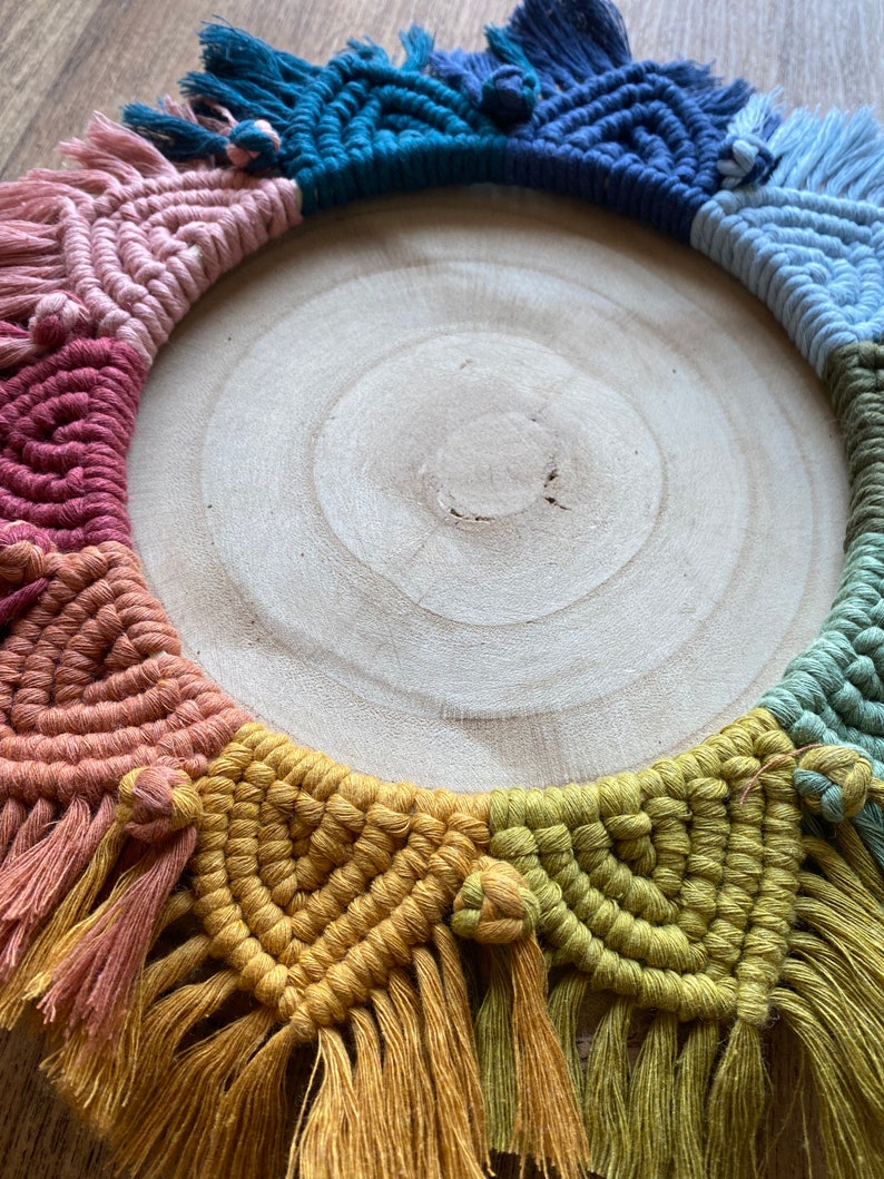 Puede incluir: Un tapiz de macram&eacute; colorido con un arco&iacute;ris de colores, incluyendo rosa, rojo, naranja, amarillo, verde, azul y morado. El macram&eacute; est&aacute; hecho con una variedad de nudos y est&aacute; terminado con borlas. El tapiz se muestra en un c&iacute;rculo de madera.