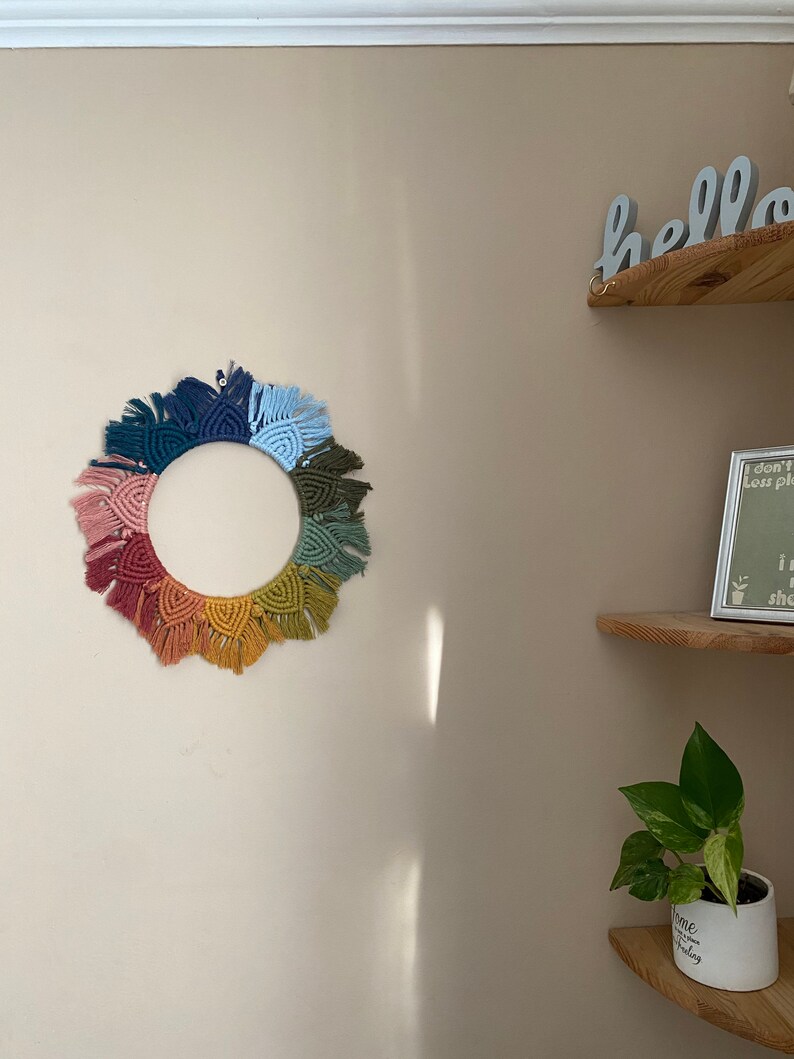 Puede incluir: Un tapiz de macram&eacute; de arco&iacute;ris colorido con forma circular y detalles de flecos. El tapiz est&aacute; hecho con diferentes tonos de hilo rosa, naranja, amarillo, verde, azul y morado.