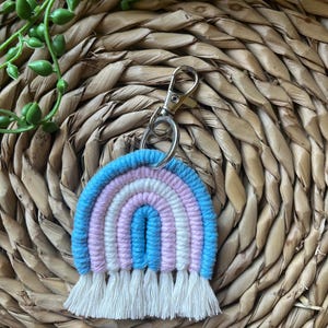 May include: A pastel-coloured macrame rainbow keyring in shades of blue, lilac, and white. It features a silver-tone clasp and a fringe of white tassels. The background is a woven, natural-toned material.