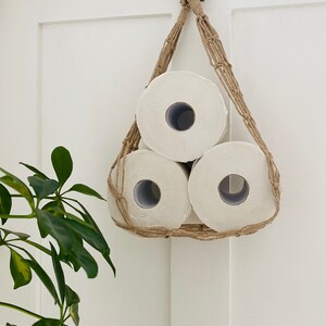 Macrame toilet paper hammock, made with taupe recycled cotton, displayed with 3 large white toilet rolls inside, against a white door with green plant next to it
