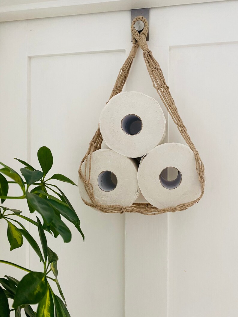 Macrame toilet paper hammock, made with taupe recycled cotton, displayed with 3 large white toilet rolls inside, against a white door with green plant next to it