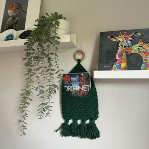 May include: A dark green macrame wall hanging with tassels, holding a book titled "Driftnet" by Fiona Maevegh. The hanging is attached to a white shelf with a wooden ring.