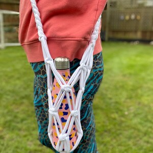 May include: White crocheted bottle holder with a pink and yellow patterned water bottle inside. The holder is worn as a crossbody bag.