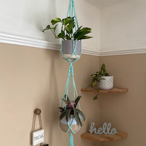 Puede incluir: Dos maceteros colgantes de macramé con cordón azul claro, uno con una planta verde y una maceta gris, el otro con una planta morada y una maceta blanca y gris. Los maceteros están colgados de un gancho de techo. La palabra "hello" está en un estante debajo de los maceteros.