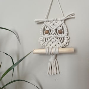 May include: A white macrame owl wall hanging with wooden dowel and brown wooden beads for eyes.