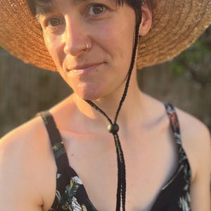 May include: A straw sun hat with a black chin strap. The hat is round with a wide brim. The person is wearing a black floral print top.
