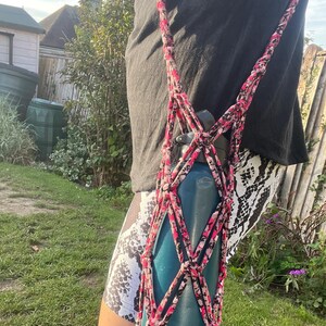 May include: A dark teal water bottle held within a handmade macrame-style carrier. The carrier is constructed from fabric strips featuring a pink, black, and white camouflage-like pattern, forming a diamond-shaped net with a long shoulder strap.