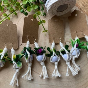 May include: Six pairs of handmade macrame earrings with small potted plants. The earrings are made with white macrame cord and feature a variety of small green and purple plants.