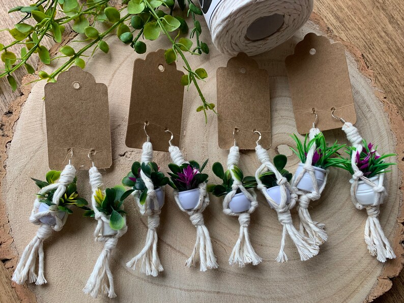 May include: Six pairs of handmade macrame earrings with small potted plants. The earrings are made with white macrame cord and feature a variety of small green and purple plants.